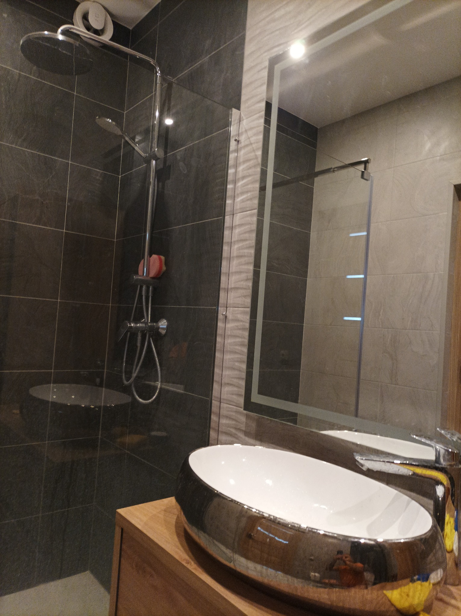 Nowoczesna łazienka z ciemnymi płytkami, deszczownicą, podświetlanym lustrem i umywalką w kształcie misy.