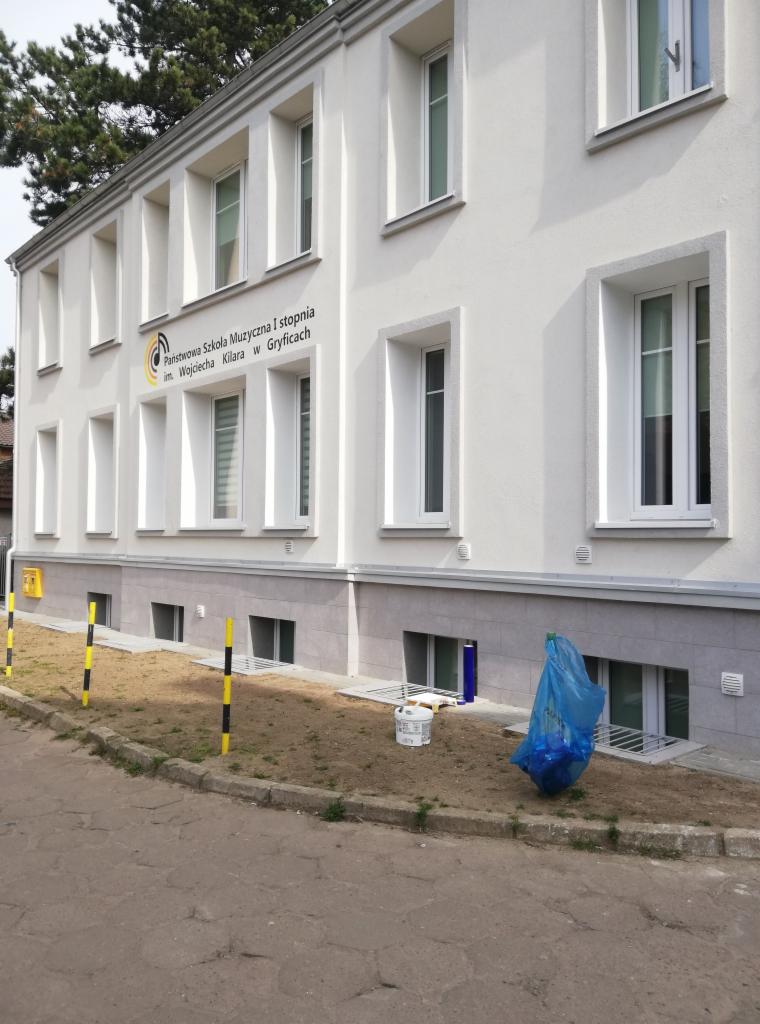 Biała elewacja budynku Państwowej Szkoły Muzycznej I stopnia im. Wojciecha Kilara w Gryficach po remoncie, z widocznymi nowymi oknami, pomalowaną fasadą, oraz elementami zabezpieczającymi teren...