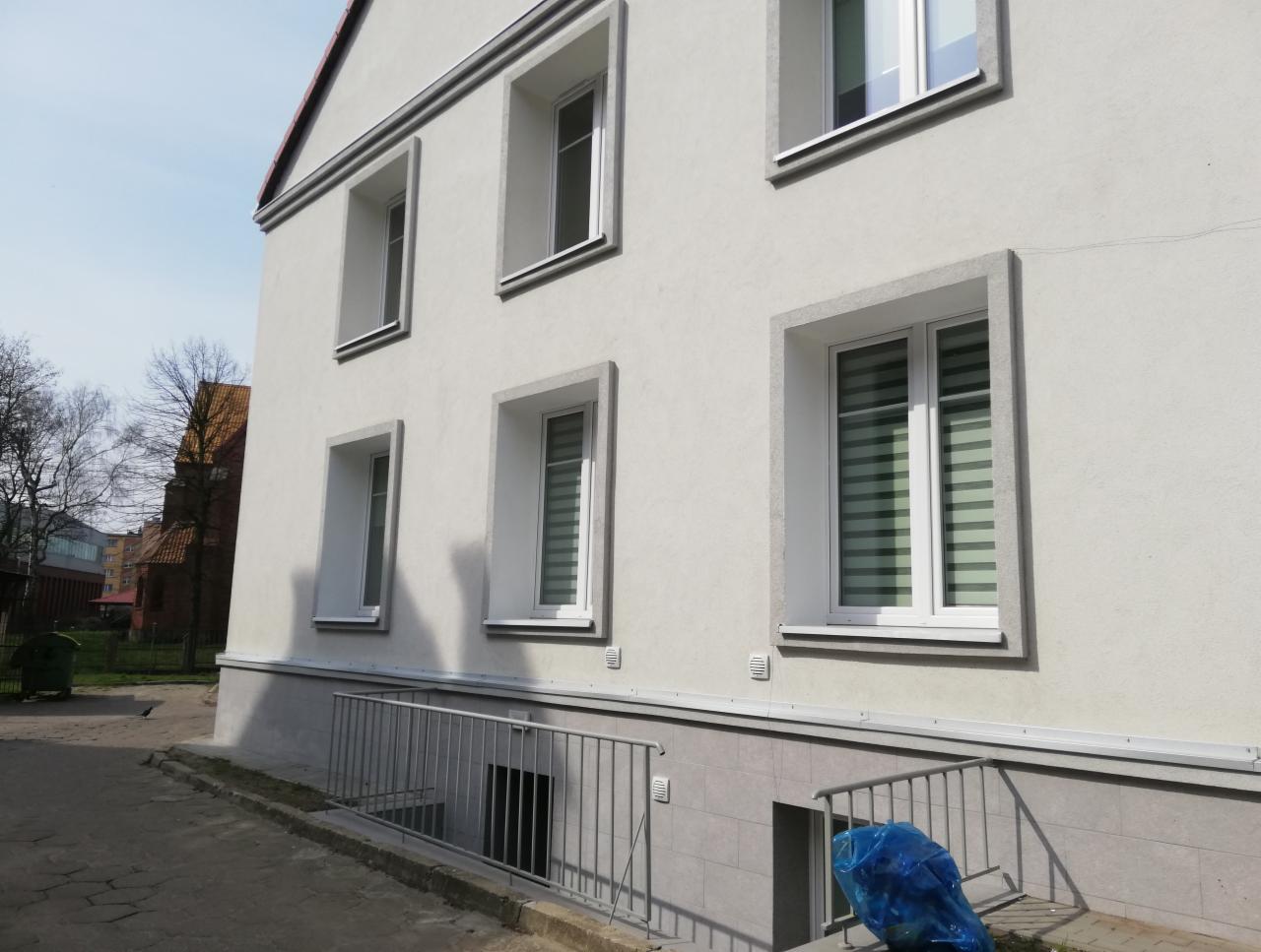 Odnowiona elewacja budynku z nowymi oknami, widoczne rolety wewnętrzne w oknach na parterze, szara podmurówka i metalowa barierka zabezpieczająca zejście do piwnicy.
