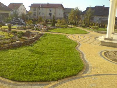 Żółta kostka brukowa tworzy kręte ścieżki w zadbanym ogrodzie z trawnikiem, skalniakiem i altaną. W tle zabudowa mieszkalna.