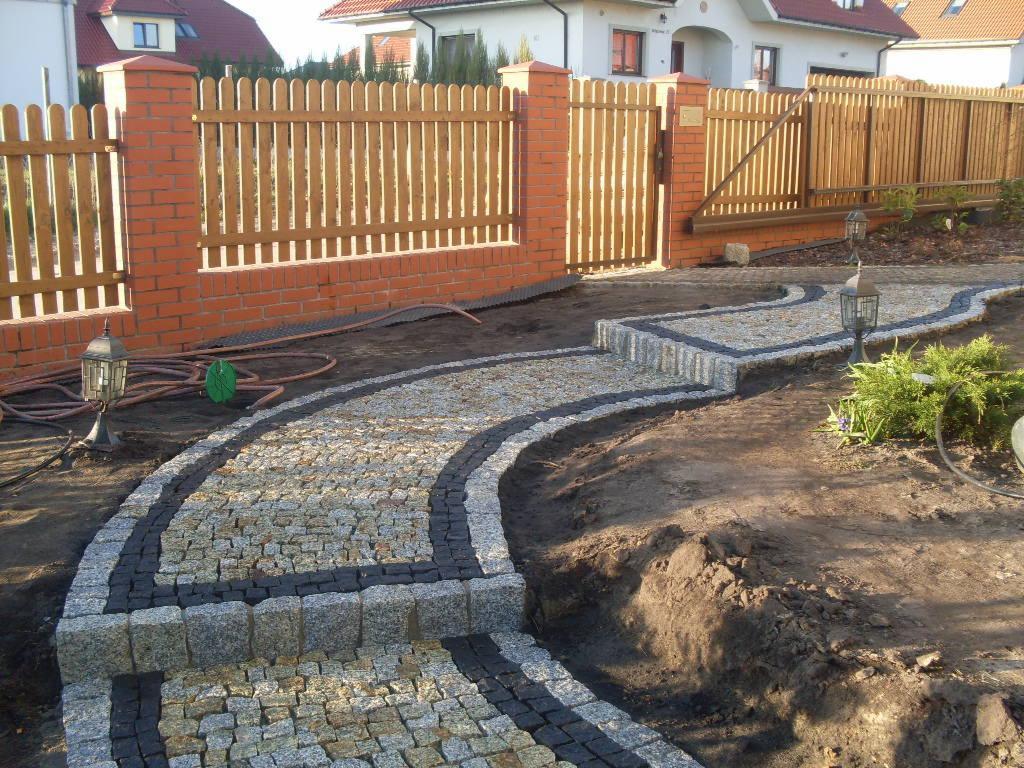 Kręta ścieżka z kostki granitowej w odcieniach szarości i czerni, obok drewnianego płotu z ceglanymi słupkami, oświetlona lampami ogrodowymi.