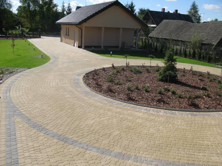 Żółta, łukowato ułożona kostka brukowa z szarymi obrzeżami tworzy podjazd do domu z ogrodem. Na środku okrągły klomb z korą i roślinami. Słoneczny dzień.