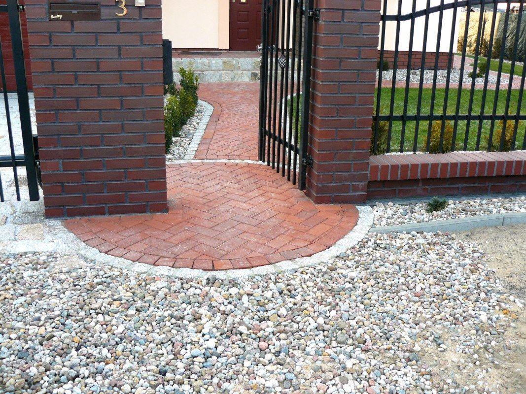 Ceglana ścieżka z kostki ułożonej w jodełkę, prowadząca przez bramę, obramowana drobnymi kamieniami. Widoczne słupki z cegły i metalowe ogrodzenie.