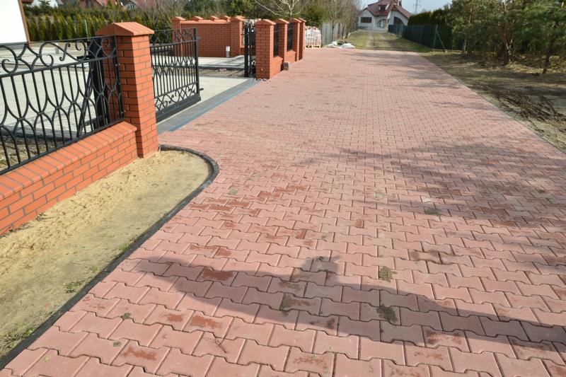 Czerwona kostka brukowa ułożona w zygzak, tworząca długi podjazd. Po lewej stronie brama z czarnym, metalowym wzorem i słupkami z czerwonej cegły.