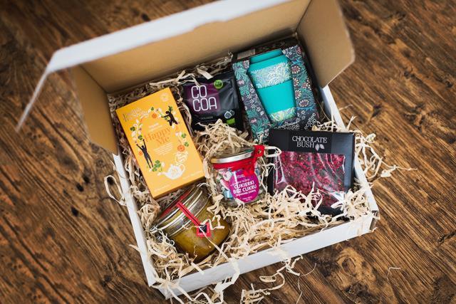 Otwarty biały karton wypełniony papierowym siankiem skrywający zestaw upominkowy: herbata Happy Me, kakao, kubek termiczny w zielono-białe wzory, słoiczek z cukierkami i czekolada z malinami...