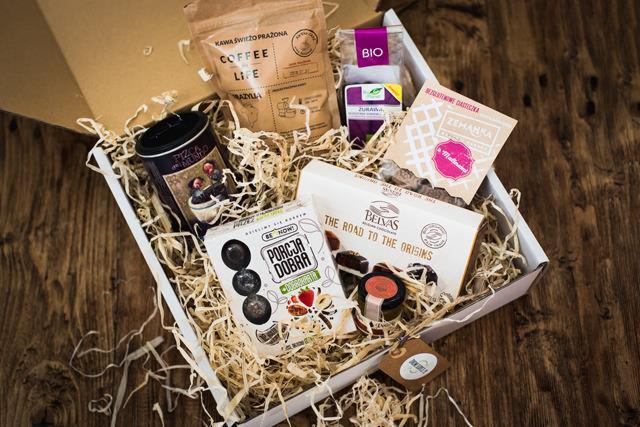 Otwarty kartonowy box wypełniony siankiem, zawierający zestaw produktów spożywczych: kawa ziarnista, herbata, czekoladki Belvas, praliny Porcja Dobra, dżem w małym słoiczku oraz słodycze Zemanka...
