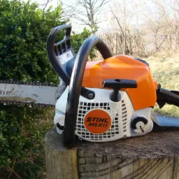 Pomarańczowo-biała pilarka spalinowa Stihl MS 211 na pniu drzewa, z zielonym krzewem i drzewami w tle.