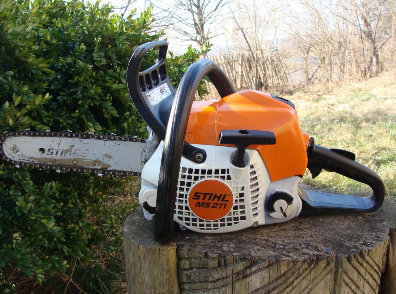 Pomarańczowo-biała pilarka spalinowa Stihl MS 211 na pniu drzewa, z zielonym krzewem i drzewami w tle.