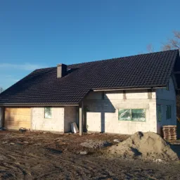 Surowy stan domu z ciemną dach&oacute;wką, okna zabezpieczone folią, widoczna sterta piasku i ślady opon w błotnistym terenie.