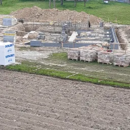 Wykop pod fundamenty z częściowo wzniesionymi ścianami z bloczków betonowych, palety z materiałem budowlanym Solbet i wykopana ziemia w tle.