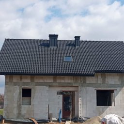 Usługi Remontowo Budowlane - Surowy dom z białego pustaka z ciemnym dachem i dwoma kominami na tle błękitnego nieba z chmurami. Widoczne otwory okienne i drzwiowe, teren budowy.
