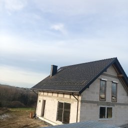 Usługi Remontowo Budowlane - Surowy dom z białego pustaka z ciemną dachówką, widoczne okna i komin. Budowa w toku, otoczenie naturalne, częściowo widoczny blaszany dach sąsiedniego budynku.