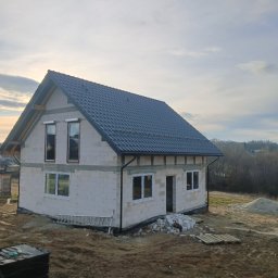 Usługi Remontowo Budowlane - Surowy dom z szarej cegły z ciemnym dachem, okna wstawione, teren wokół niezagospodarowany, drabina oparta o ścianę, widok na zewnątrz z lekkiego wzniesienia.