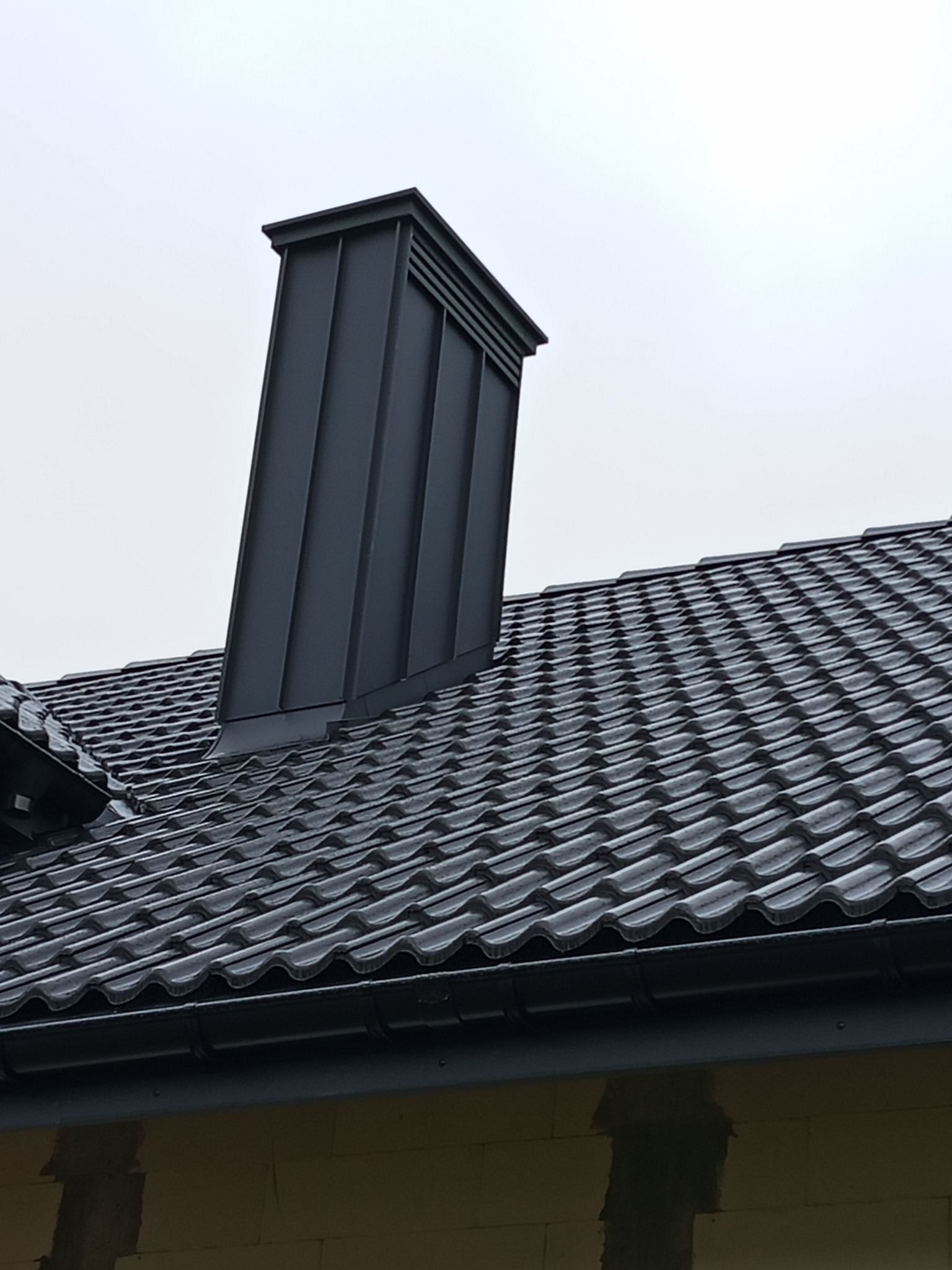 Szary komin z blachy na dachu z ciemnej dachówki ceramicznej. Widoczne orynnowanie i fragment ściany budynku z pustaków. Pochmurne niebo w tle.