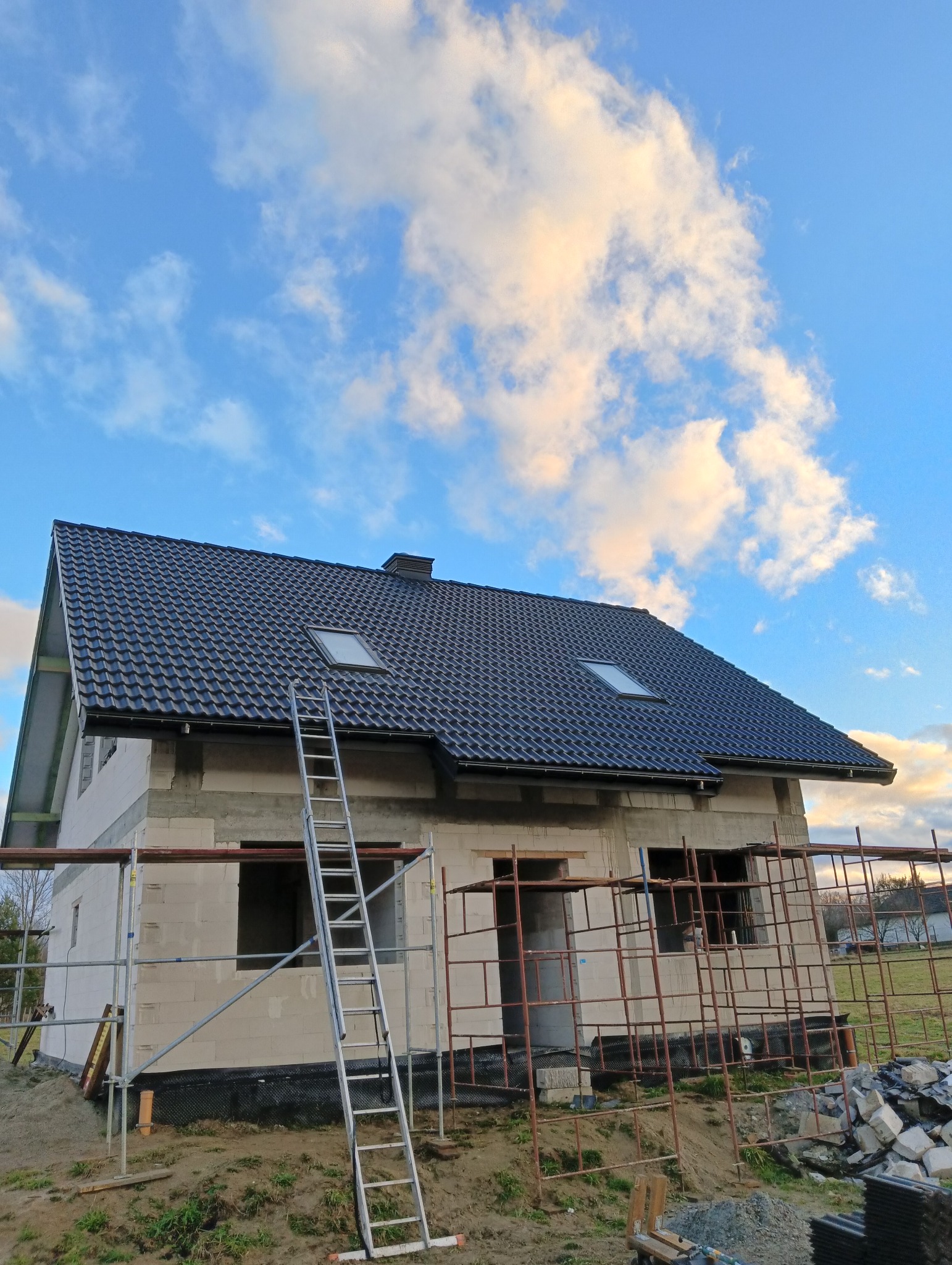 Dom w stanie surowym zamkniętym z czarną dachówką i oknami dachowymi w Zagórzanach. Widoczne rusztowania i drabina oparta o budynek. Błękitne niebo z chmurami w tle.