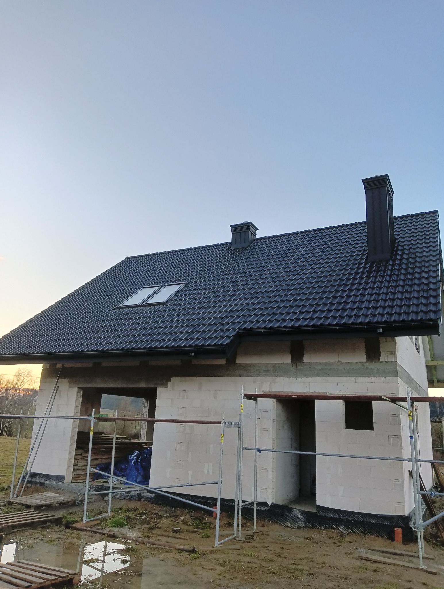 Dom w trakcie budowy z czarnym dachem i dwoma kominami. Widoczne rusztowania i materiały budowlane. Częściowo wykończony dach z oknami dachowymi.