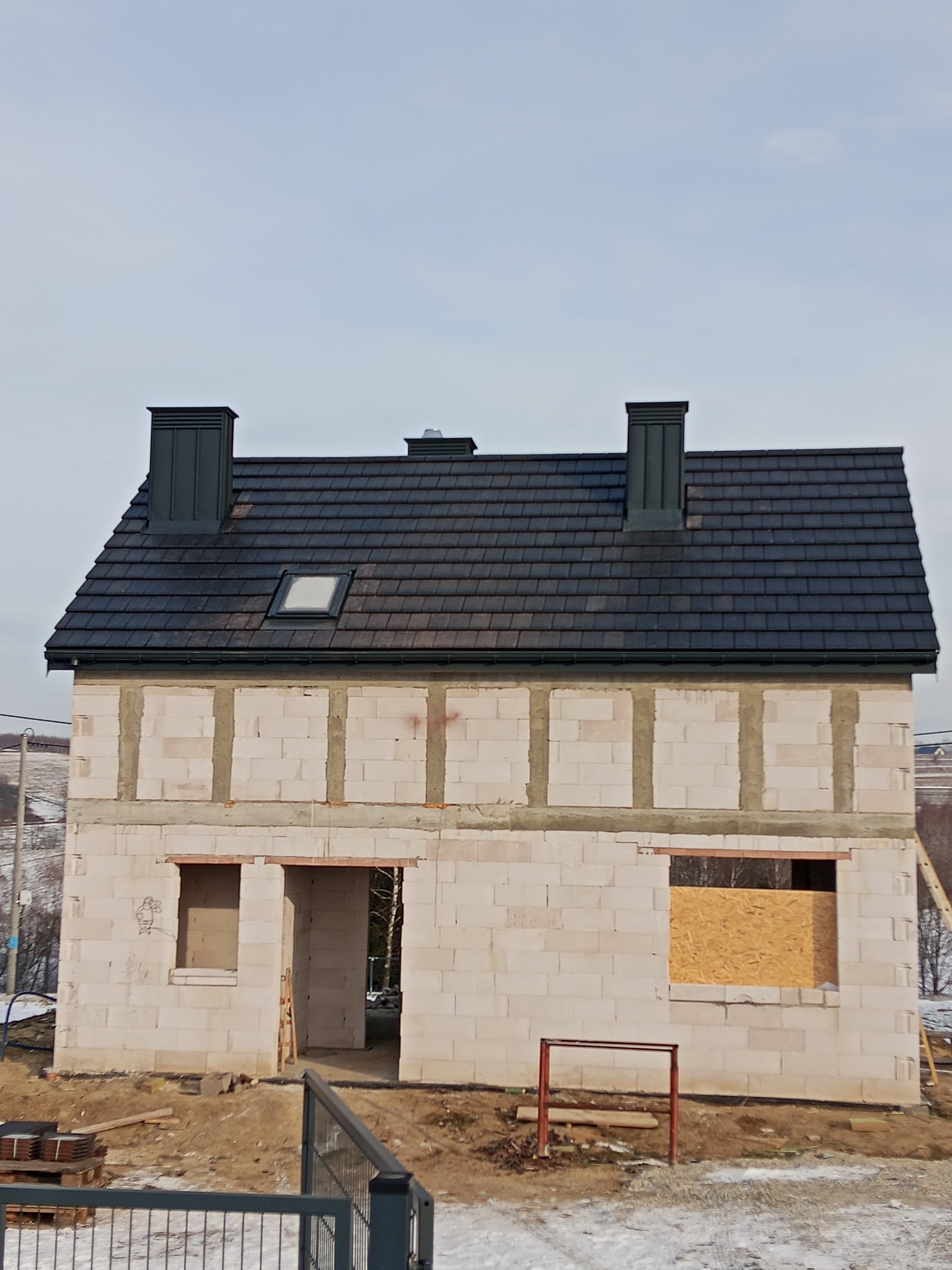 Niedokończony dom z białych bloczków, z ciemnym dachem i dwoma kominami. Okna częściowo zabite deskami, widoczne elementy rusztowania i otoczenie budowy.