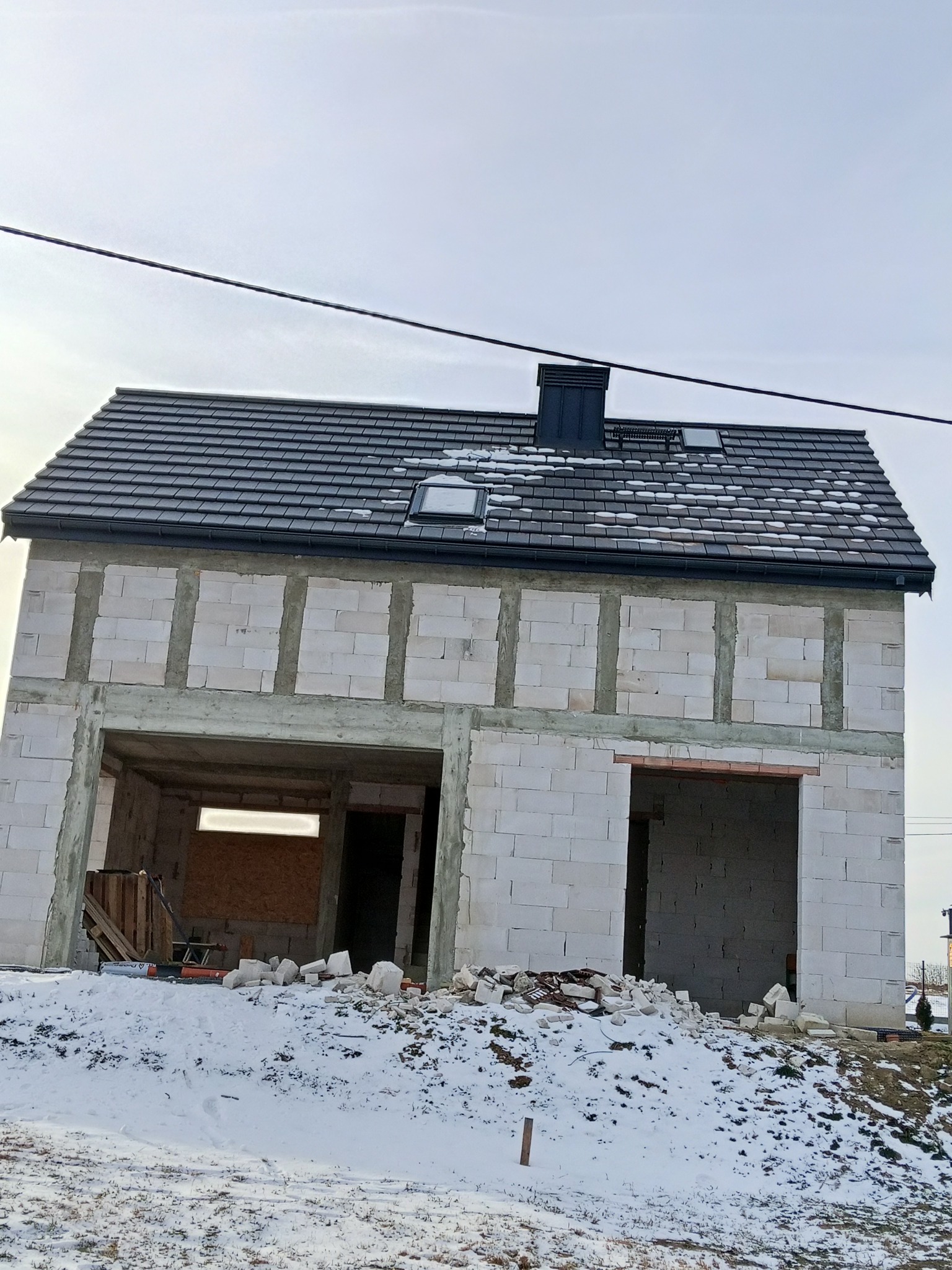 Niedokończony, dwukondygnacyjny budynek z białych bloczków, z ciemnym dachem i kominem, w zimowym krajobrazie. Widoczne otwory okienne i drzwiowe, śnieg na dachu i ziemi.