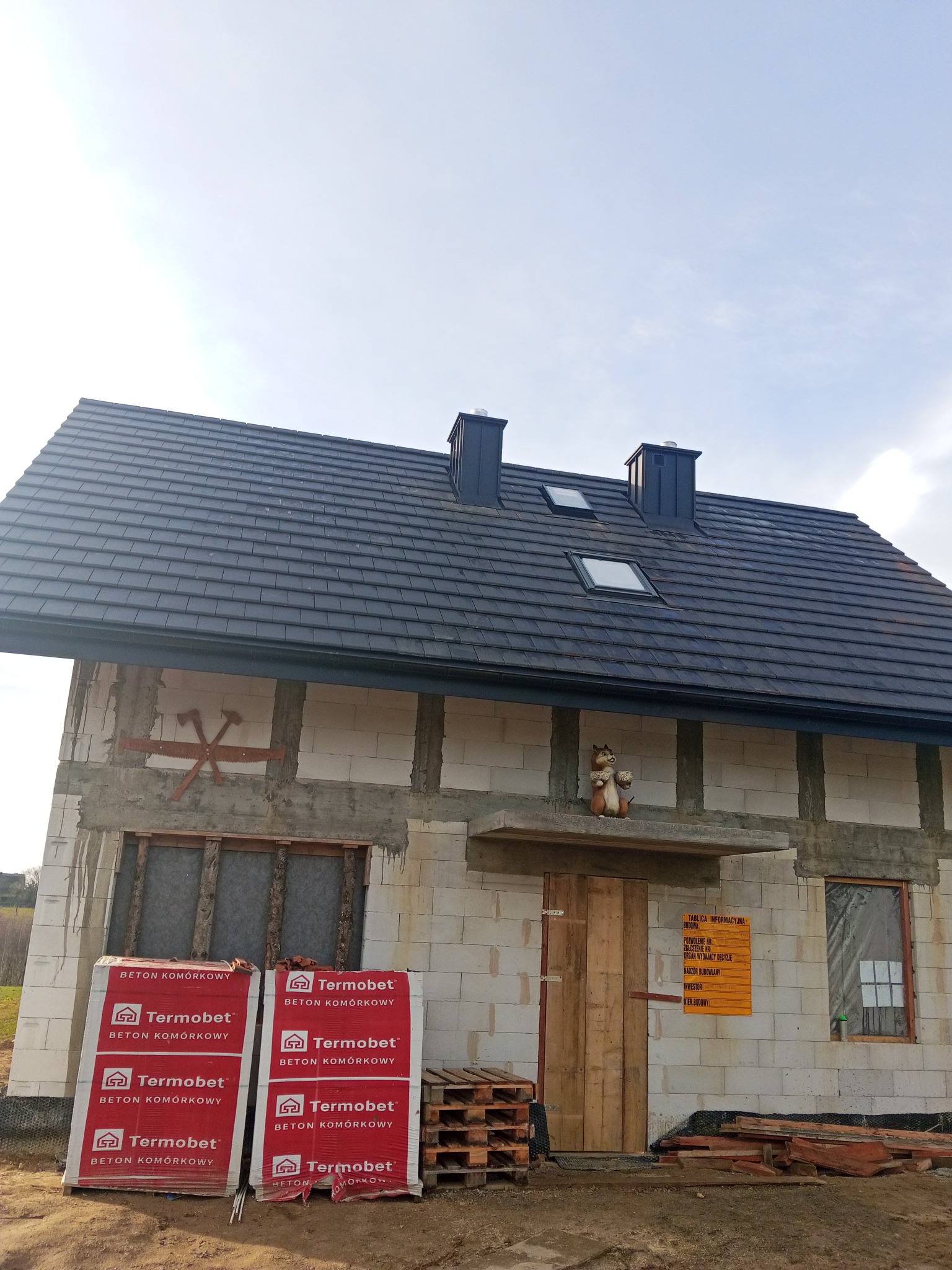 Niedokończony dom z betonu komórkowego Termobet, czarnym dachem z oknami połaciowymi i dwoma kominami. Na ścianie tablica informacyjna budowy i dekoracja.