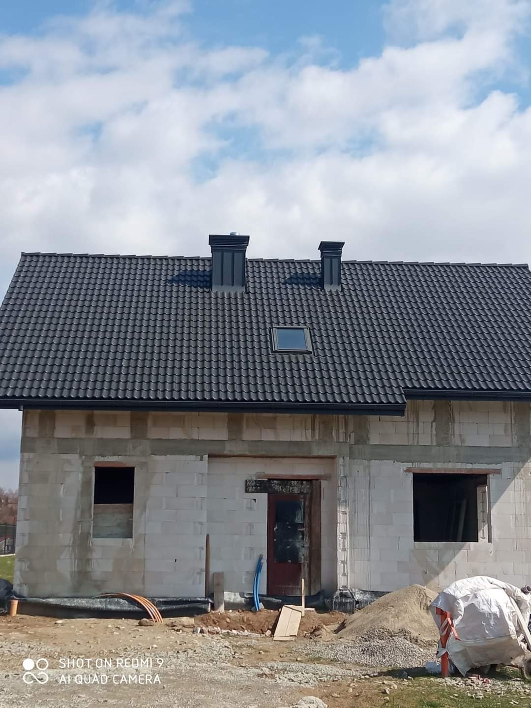 Surowy dom z białego pustaka z ciemnym dachem i dwoma kominami na tle błękitnego nieba z chmurami. Widoczne otwory okienne i drzwiowe, teren budowy.