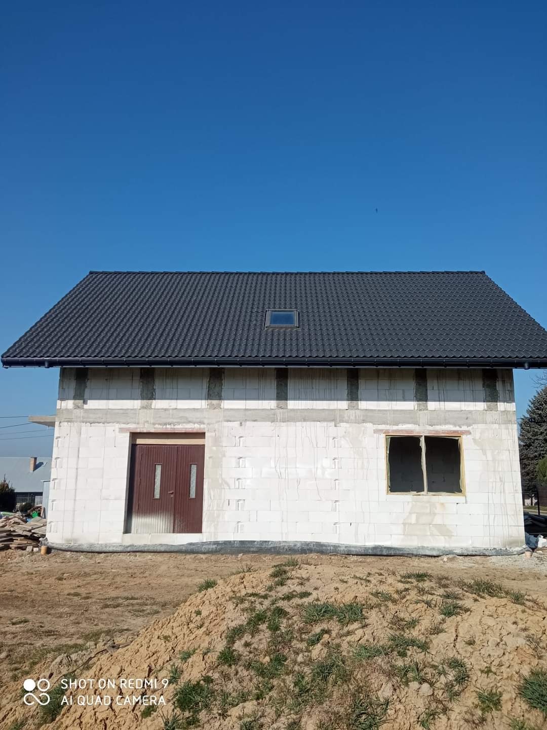 Surowy dom jednorodzinny z ciemnym dachem i oknem dachowym, w trakcie budowy. Widoczne białe bloczki, otwory okienne i drzwiowe. Błękitne niebo w tle.