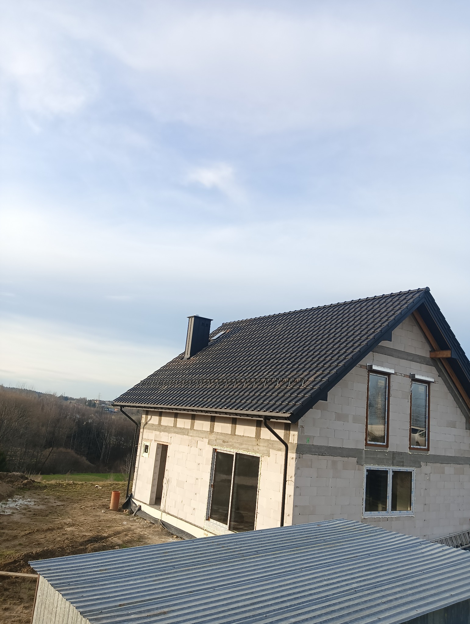 Surowy dom z białego pustaka z ciemną dachówką, widoczne okna i komin. Budowa w toku, otoczenie naturalne, częściowo widoczny blaszany dach sąsiedniego budynku.