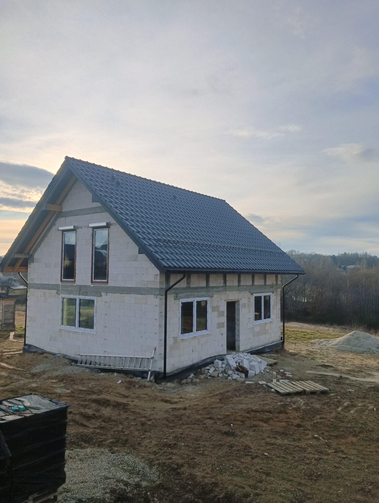 Surowy dom z szarej cegły z ciemnym dachem, okna wstawione, teren wokół niezagospodarowany, drabina oparta o ścianę, widok na zewnątrz z lekkiego wzniesienia.