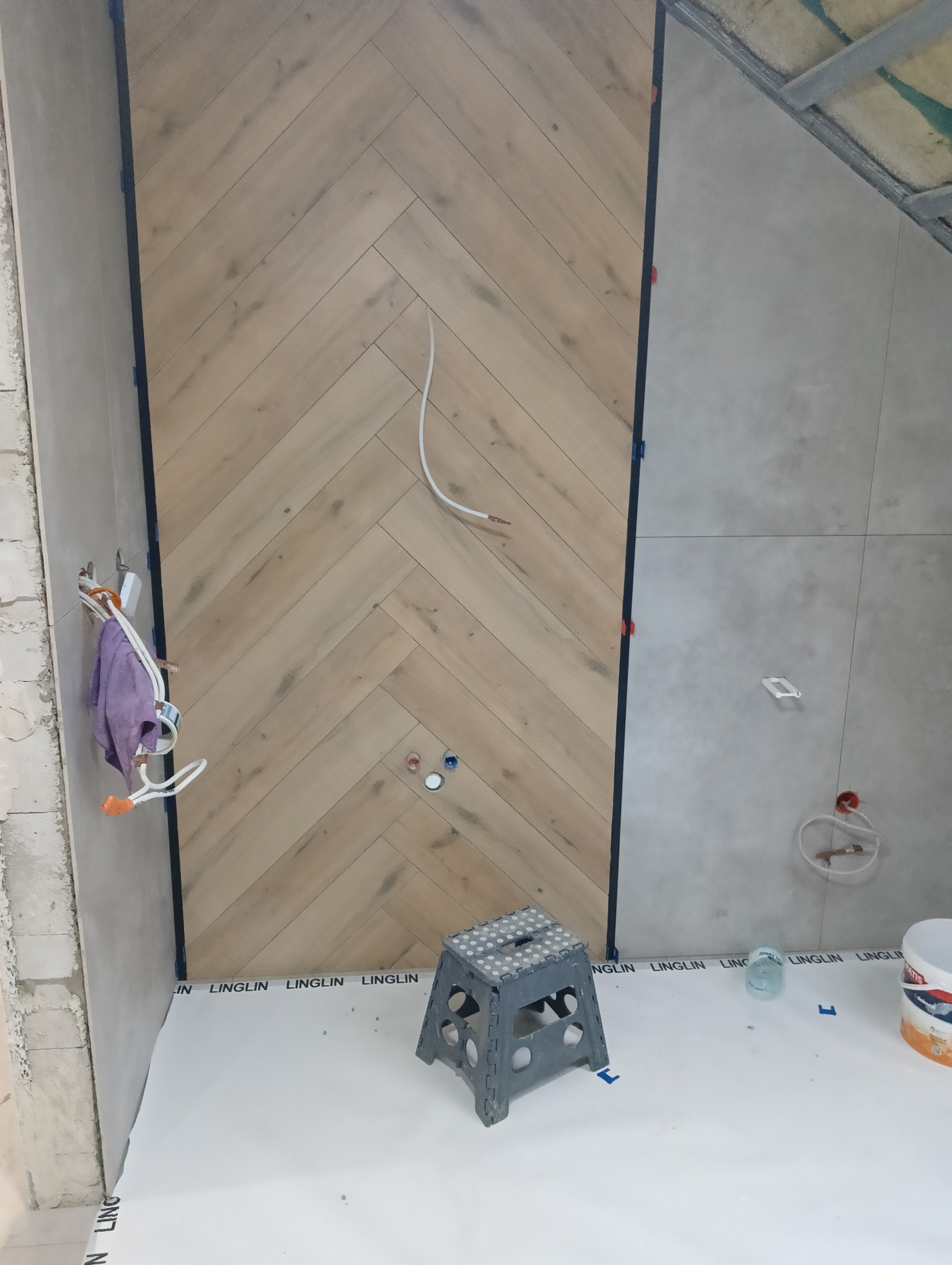 Ściana wyłożona drewnopodobnymi płytkami w jodełkę, fragment ściany z szarymi płytkami, instalacja elektryczna i drabinka. Prace wykończeniowe w łazience.