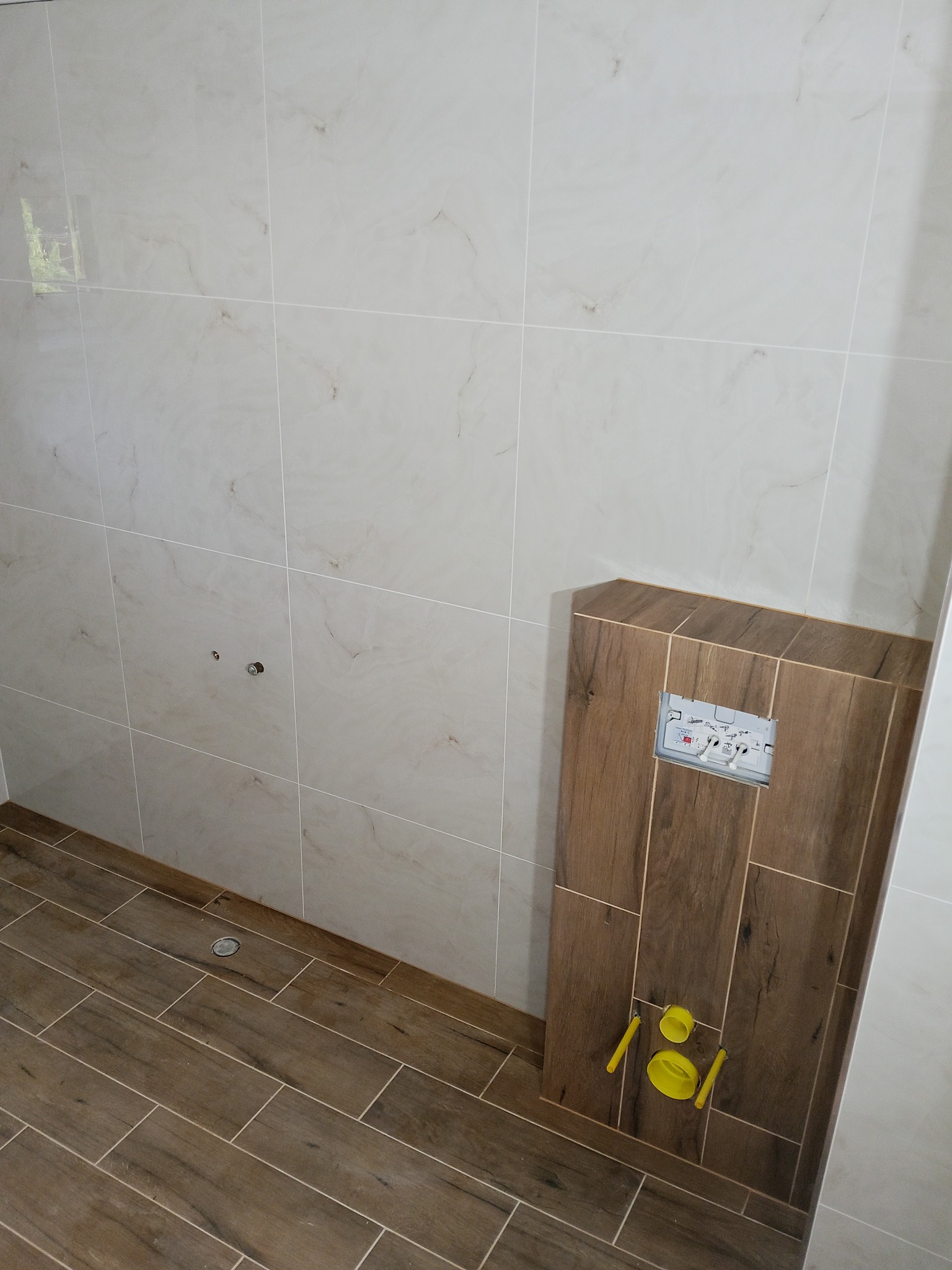 Wykończona łazienka z zabudową stelaża WC w płytkach imitujących drewno, kremowe płytki ścienne z delikatnym wzorem i brązowa podłoga z płytek. Widoczny odpływ liniowy.