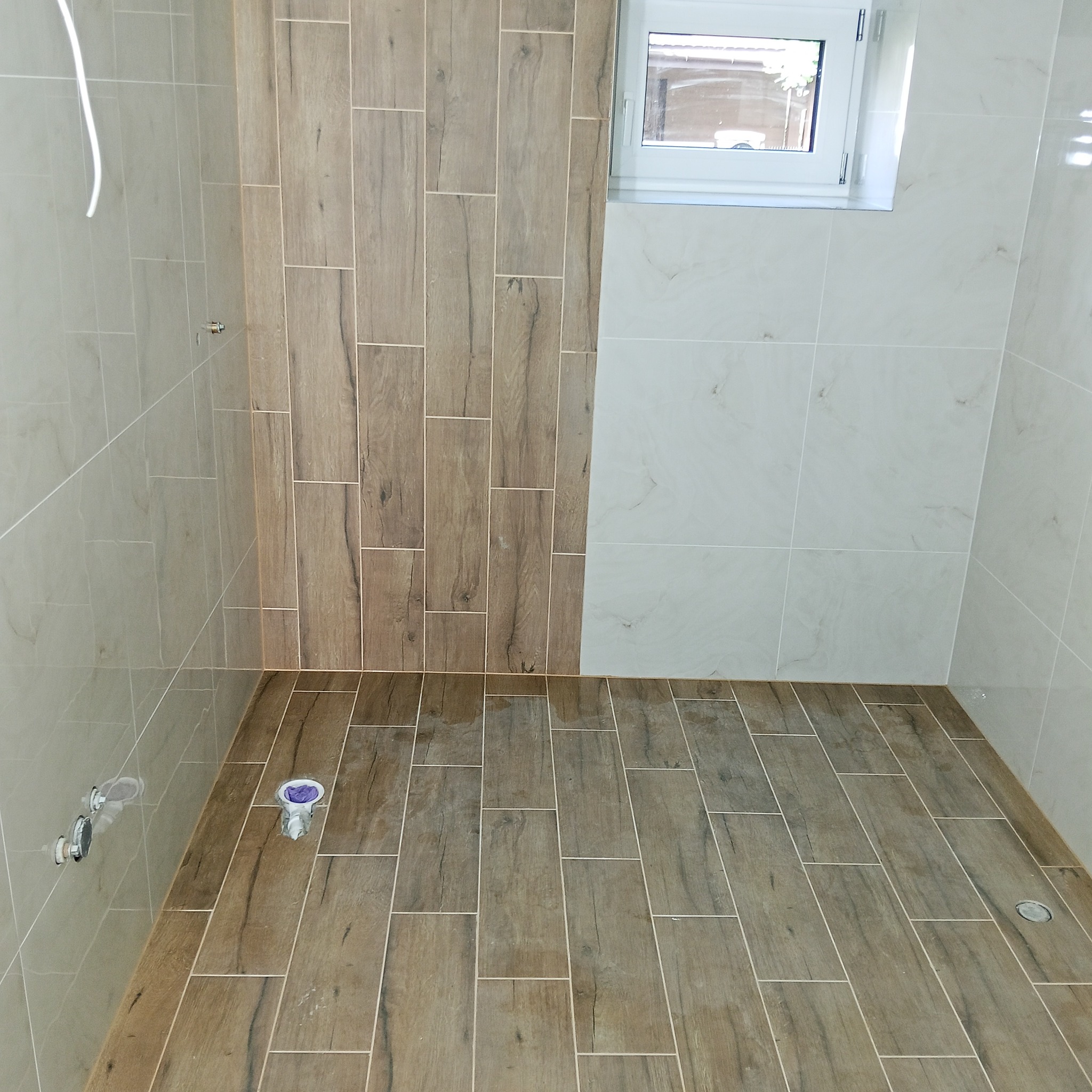 Wykończona łazienka z drewnopodobnymi płytkami na podłodze i ścianie, kontrastującymi z jasnymi płytkami. Widoczne przyłącza wodne i okno.