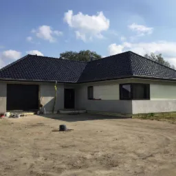 Niedokończony parterowy dom z garażem, czarne dachówki na dachu, ściany częściowo ocieplone, widoczne materiały budowlane na podjeździe.
