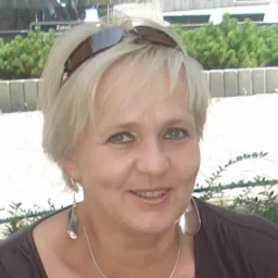 Portret uśmiechniętej kobiety z blond włosami, okularami na głowie i srebrną biżuterią na tle zieleni.