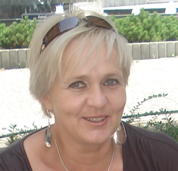 Portret uśmiechniętej kobiety z blond włosami, okularami na głowie i srebrną biżuterią na tle zieleni.