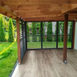 Zadaszona konstrukcja z drewnianym stropem i podłogą imitującą drewno, z ażurowymi ścianami bocznymi, widok na trawnik i tuje w tle.