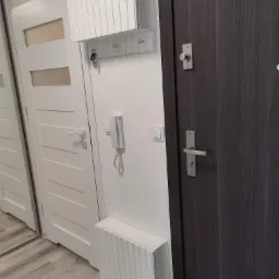 Przedpokój z białymi drzwiami z przeszkleniami, ciemnymi drzwiami z metalową klamką, białym grzejnikiem dekoracyjnym z pionowymi żłobieniami oraz szafką na klucze z wieszakami i interkomem na ścianie.