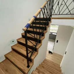 Wnętrze domu w trakcie wykańczania: drewniane schody z czarną metalową balustradą zabezpieczoną niebieską taśmą, widoczne ślady po pracach na ścianie, w tle fragment kuchni.