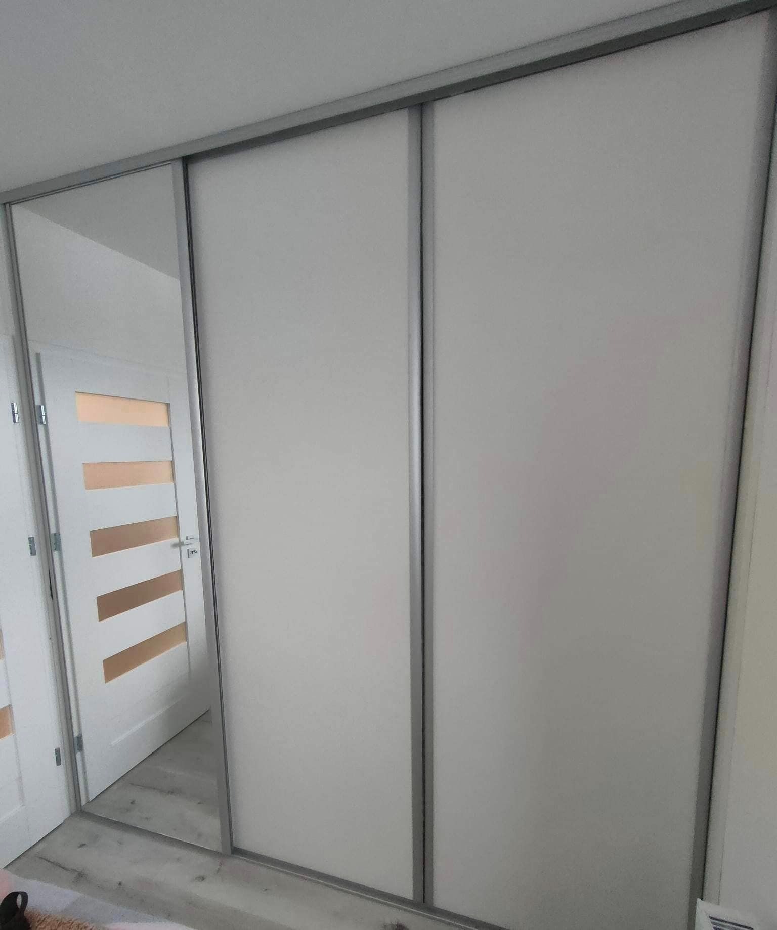 Trzydrzwiowa szafa wnękowa z przesuwnymi drzwiami w kolorze białym i srebrnymi obramowaniami, z lustrzanym frontem po lewej stronie, odbijającym białe drzwi z poziomymi przeszkleniami.