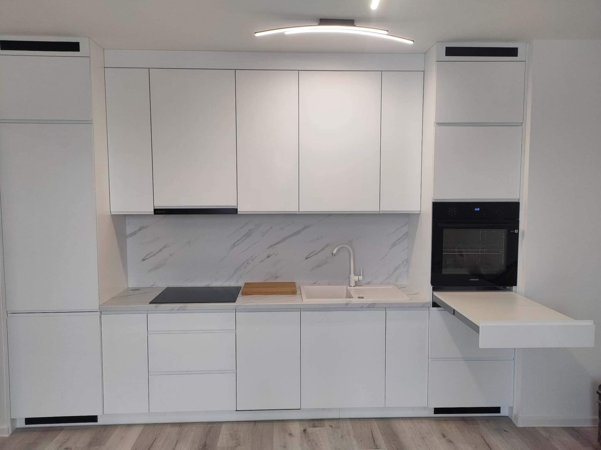 Nowoczesna, minimalistyczna kuchnia w bieli z marmurowym akcentem na ścianie, wyposażona w płytę indukcyjną, piekarnik i rozkładany blat roboczy, oświetlona ledowym plafonem.