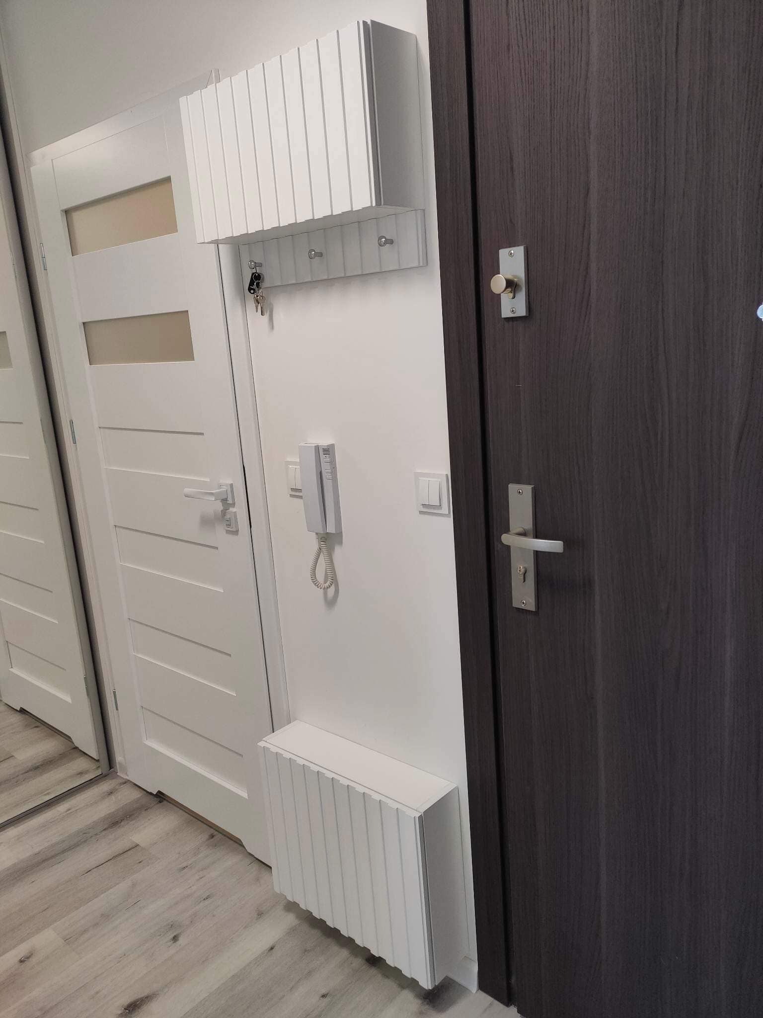 Przedpokój z białymi drzwiami z przeszkleniami, ciemnymi drzwiami z metalową klamką, białym grzejnikiem dekoracyjnym z pionowymi żłobieniami oraz szafką na klucze z wieszakami i interkomem na ścianie.