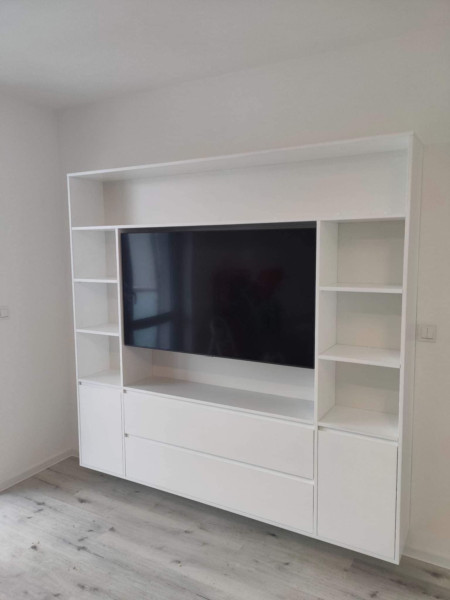 Biała, nowoczesna zabudowa meblowa z miejscem na telewizor, szufladami i półkami, na tle jasnej ściany i podłogi imitującej drewno.