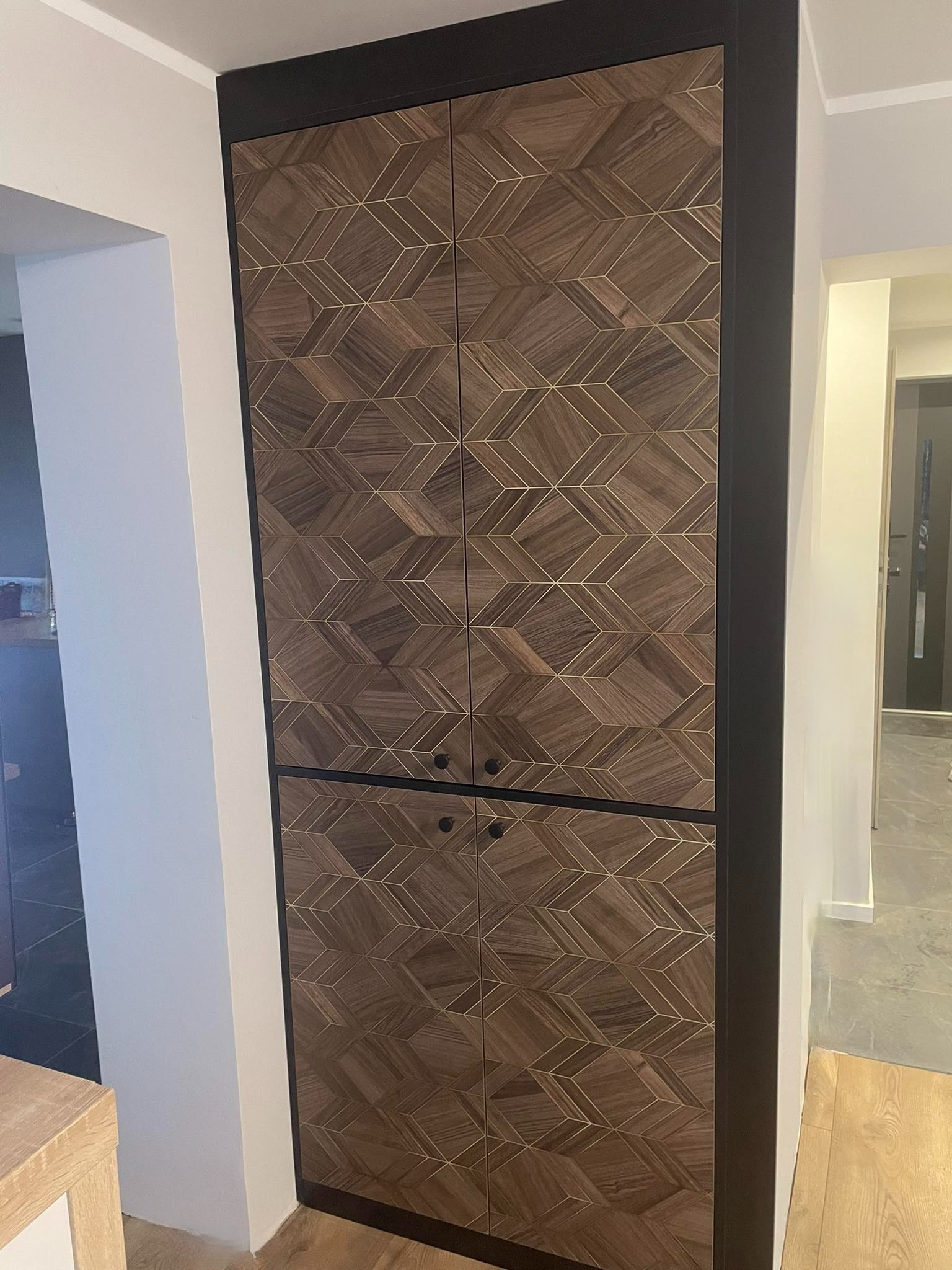Wysoka szafa wnękowa z drewnianymi frontami o geometrycznym wzorze z motywem sześcianów i złotych linii, czarne obramowanie, widok z boku na korytarz.