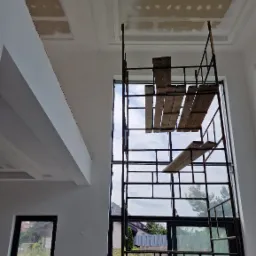 Wysokie wnętrze w trakcie prac wykończeniowych: sufit z płyt kartonowo-gipsowych częściowo wyszpachlowany, rusztowanie przed dużym oknem, widok na zewnątrz.