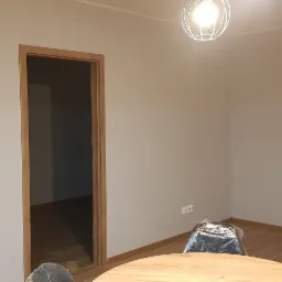 Jasne wnętrze mieszkania po remoncie, z pomalowanymi ścianami w odcieniach szarości, drewnianą stolarką drzwiową i oknami balkonowymi z widokiem na miasto w tle. Na podłodze panele, widoczny...