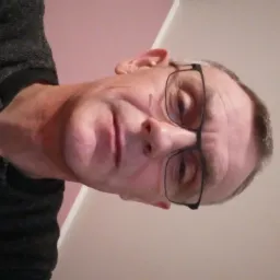 Portret mężczyzny w okularach, z bliska, na tle ściany w odcieniach beżu i różu. Ubrany w ciemną koszulę. Zdjęcie obrócone w lewo.