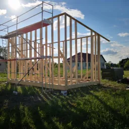 Wznoszenie szkieletu drewnianego budynku, widoczne rusztowanie z czerwonym elementem, w tle domy jednorodzinne i błękitne niebo z promieniami słońca.