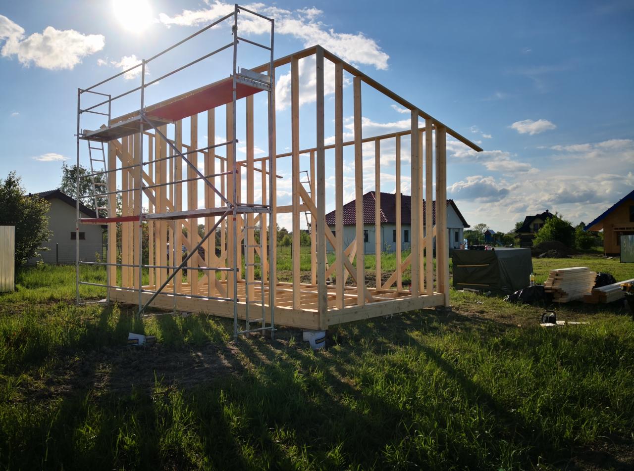 Wznoszenie szkieletu drewnianego budynku, widoczne rusztowanie z czerwonym elementem, w tle domy jednorodzinne i błękitne niebo z promieniami słońca.