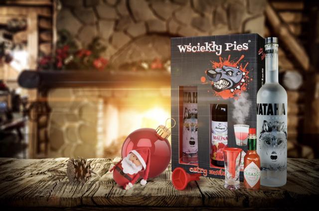 Świąteczny zestaw prezentowy z wódką, ostrym sosem i akcesoriami do drinków na drewnianym stole, w tle kominek i dekoracje świąteczne.