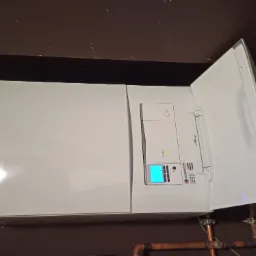 Biały, wiszący kocioł gazowy z otwartą obudową, widoczne miedziane rury instalacji grzewczej i panel sterowania z niebieskim wyświetlaczem, zamontowany pod ciemnym sufitem.