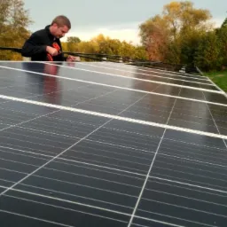 Mężczyzna w czarnej kurtce montuje panele słoneczne na dachu, używając czerwonej wkrętarki; widoczny krajobraz z drzewami w tle.