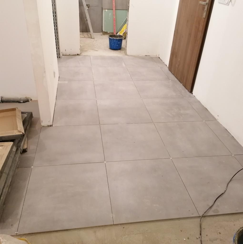 Ułożona szara podłoga z płytek gresowych w trakcie prac wykończeniowych, widoczne krzyżyki dystansowe, częściowo odsłonięta surowa podłoga, wiadro i drabina w tle.